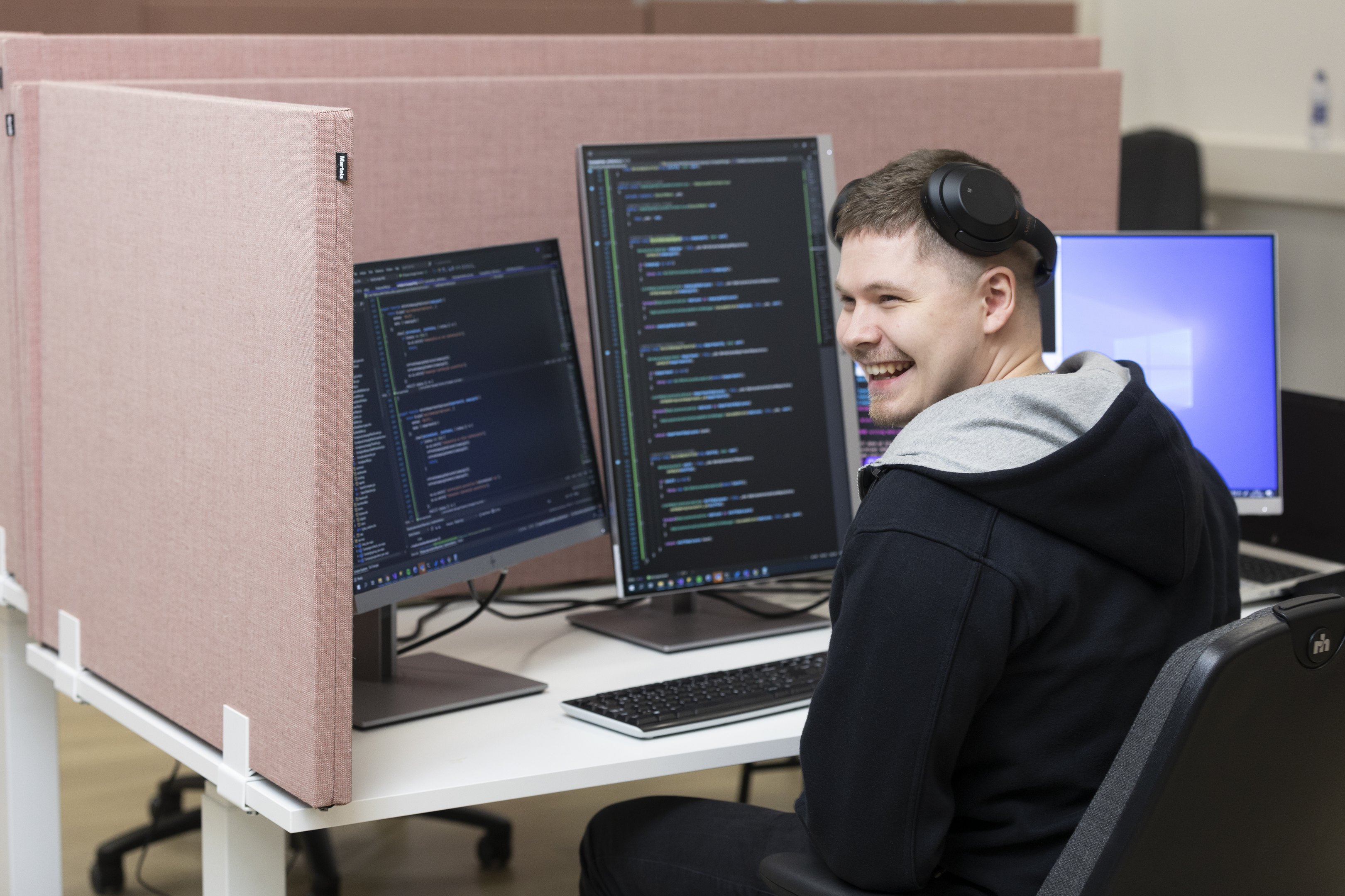 Hymyilevä ohjelmistokehittäjä työpisteellä, edessä koodia näytöillä